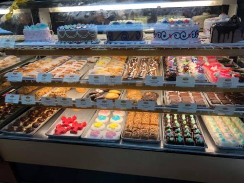 A display case filled with various colorful cakes, pastries, and desserts, labeled with names and prices.