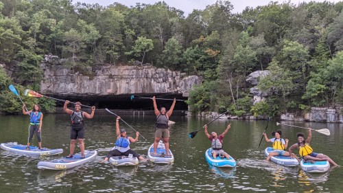 A group of people on paddleboards in a lake, celebrating with raised arms, surrounded by lush greenery and rocky cliffs.