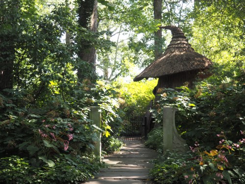 A stone pathway leads through lush greenery to a whimsical thatched-roof structure in a serene garden.