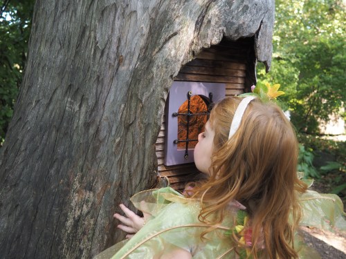 A child in a fairy costume leans toward a small wooden door in a tree, peering inside with curiosity.