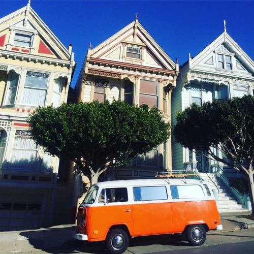 Colorful Victorian houses with a bright orange van parked in front, surrounded by lush green trees.