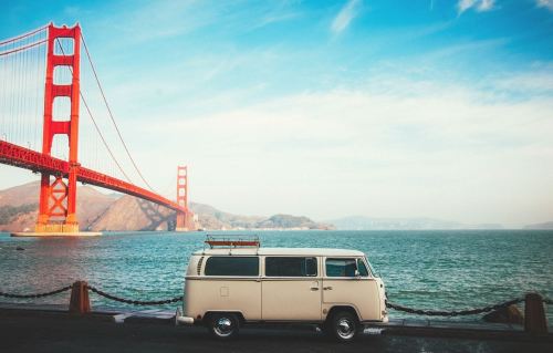 A vintage van parked by the water with the Golden Gate Bridge in the background under a clear blue sky.