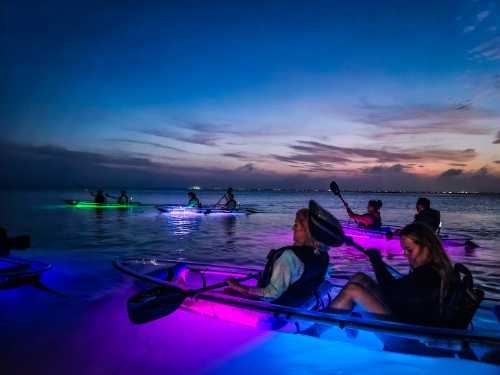 A group of kayakers paddles at dusk, illuminated by colorful lights on their kayaks, reflecting on the water.