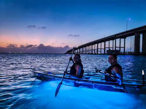 Two women kayak in glowing water under a bridge at dusk, with a colorful sky in the background.