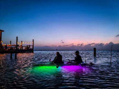 Two people in a kayak with colorful lights, paddling on calm water at sunset, with a pier in the background.