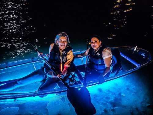 Two women in a brightly lit kayak at night, smiling and holding paddles, with shimmering water around them.