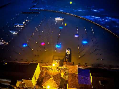 Aerial view of a harbor at night, featuring boats and illuminated umbrellas in the water, with waves in the background.