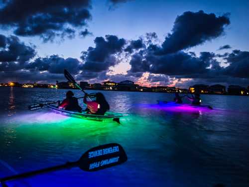 Two kayakers paddle in a glowing, colorful waterway at dusk, with clouds and lights from nearby buildings in the background.