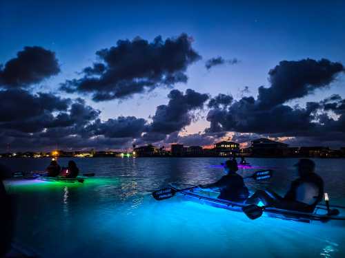 Kayakers with glowing lights paddle on a calm waterway at dusk, surrounded by clouds and city lights.