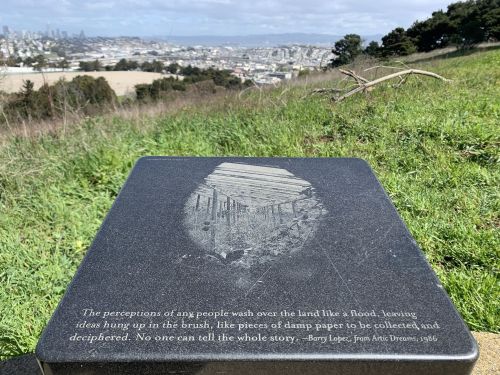 A stone plaque with an engraved quote by Barry Lopez, overlooking a cityscape and green hills.