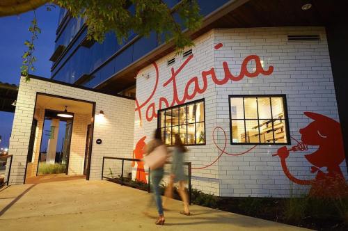 A modern restaurant exterior with a large "Pastaia" mural, featuring two people walking by at dusk.