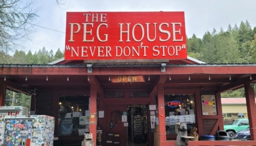 A red building with a sign reading "The Peg House" and "Never Don't Stop," surrounded by trees. Open sign visible.