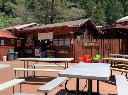 A rustic outdoor food stand with picnic tables, surrounded by trees, offering burgers and lemonade.