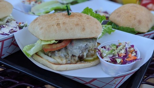 A close-up of a juicy burger with lettuce, tomato, and cheese, served with a side of coleslaw in a small cup.