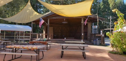 Outdoor stage area with picnic tables, shaded by yellow canopies, surrounded by trees and American flags.