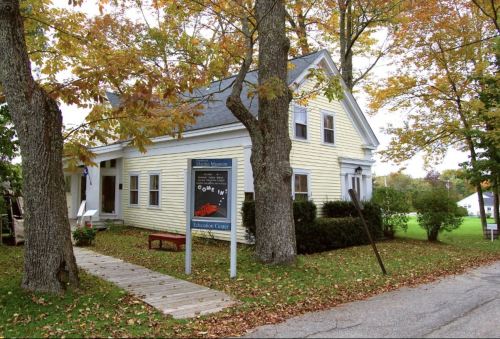 A yellow house with a sign in front, surrounded by trees with autumn leaves, on a quiet street.