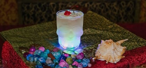 A glowing skull-shaped glass filled with a cocktail, topped with a cherry, surrounded by colorful stones and a seashell.