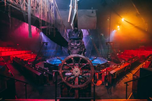 A dimly lit circus arena featuring a ship's wheel and a pirate-themed backdrop, with colorful lights and empty seating.