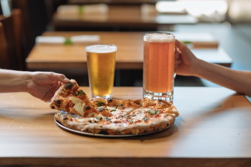 A hand reaches for a slice of pizza on a table, with two glasses of beer nearby.