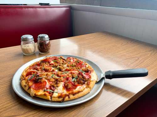 A freshly baked pizza topped with pepperoni, sausage, and vegetables on a silver platter, with condiments in the background.