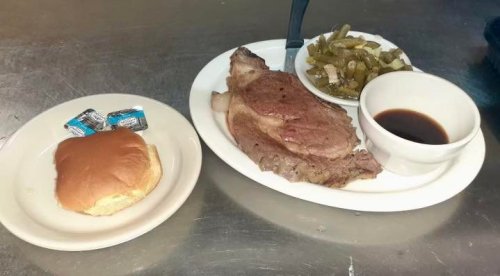 A plate with a cooked steak, green beans, a small bowl of sauce, and a bun with butter packets on the side.
