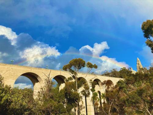 A stone viaduct arches under a vibrant rainbow against a blue sky with fluffy clouds and trees in the foreground.