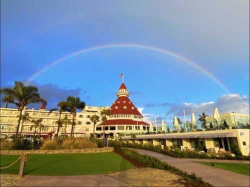 A colorful rainbow arcs over a large, red-roofed hotel surrounded by palm trees and blue skies.