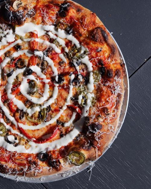 A close-up of a pizza topped with olives, peppers, and a swirl of white sauce on a dark surface.