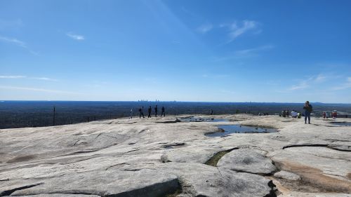 A panoramic view from a rocky summit, with people silhouetted against a clear blue sky and distant horizon.