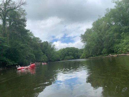 A serene river scene with lush green trees, cloudy skies, and people floating on red rafts. Reflections on the water.