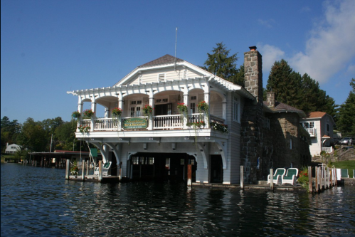 A charming lakeside house with a stone chimney, balconies, and flower boxes, surrounded by water and trees.
