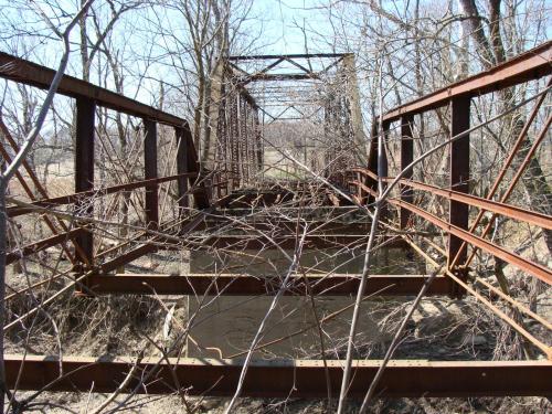 An old, rusty metal bridge over a narrow stream, surrounded by bare trees and overgrown branches.