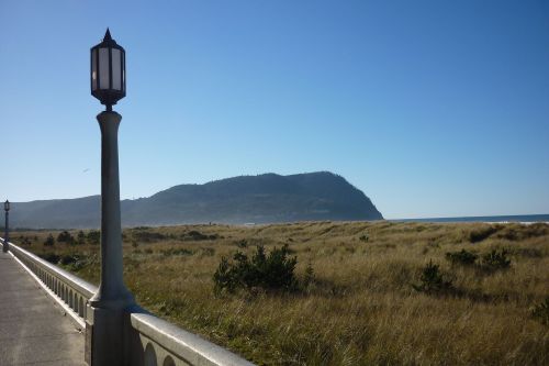 A coastal view featuring a pathway, a lamp post, and a grassy area leading to a mountain in the distance under a clear sky.
