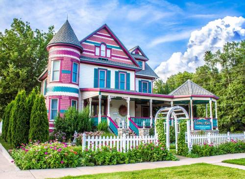 A colorful Victorian-style house with a turret, surrounded by lush greenery and vibrant flowers, under a blue sky.