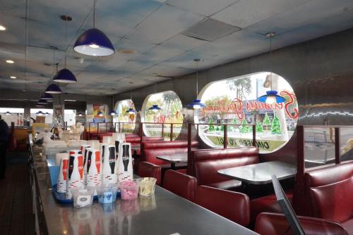 A retro diner interior with red booths, a shiny metal counter, and colorful drink cups, featuring large windows.