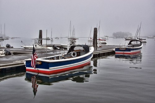 Foggy harbor scene with boats docked, reflecting in calm water, and a muted atmosphere.