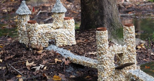 A miniature castle made of pebbles and stones, with a bridge, set in a wooded area surrounded by fallen leaves.