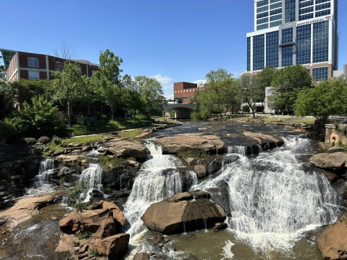 A scenic view of a waterfall cascading over rocks, with greenery and a modern building in the background.