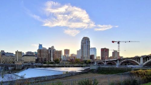 City skyline at sunset with a river in the foreground and a construction crane in the background.