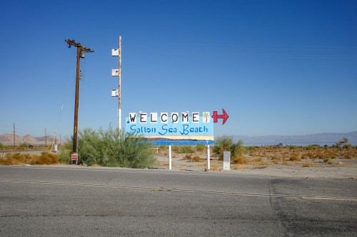A colorful "Welcome to Salton Sea Beach" sign beside a road, with desert landscape and clear blue sky in the background.