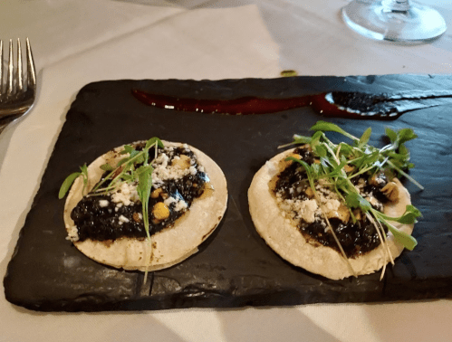 Two small tortillas topped with a dark sauce, crumbled cheese, and fresh greens, served on a black slate plate.