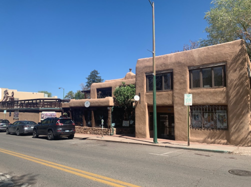 A street view of adobe-style buildings with a clear blue sky, featuring parked cars and a pedestrian bridge in the background.