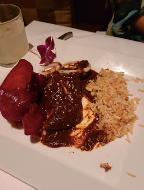 A plate of mole sauce with chicken, rice, and a side of red peppers, garnished with an orchid.