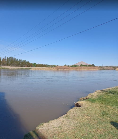 A calm river flows beside grassy banks under a clear blue sky, with distant mountains in the background.