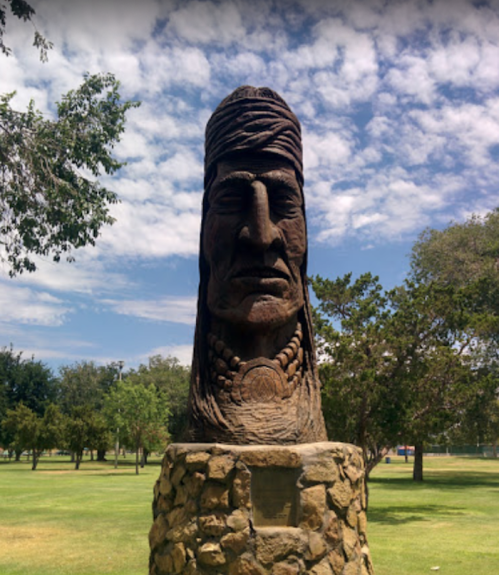 A large stone sculpture of a Native American figure stands in a grassy park under a blue sky with scattered clouds.
