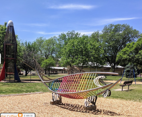 Colorful fish-shaped sculpture in a park, surrounded by trees and playground equipment under a clear blue sky.