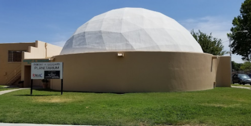 A large, white geodesic dome structure on a grassy area, with a sign for a planetarium nearby.