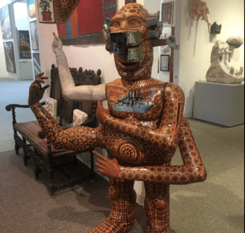 A large, intricately patterned sculpture of a humanoid figure with one arm raised, displayed in an art gallery.