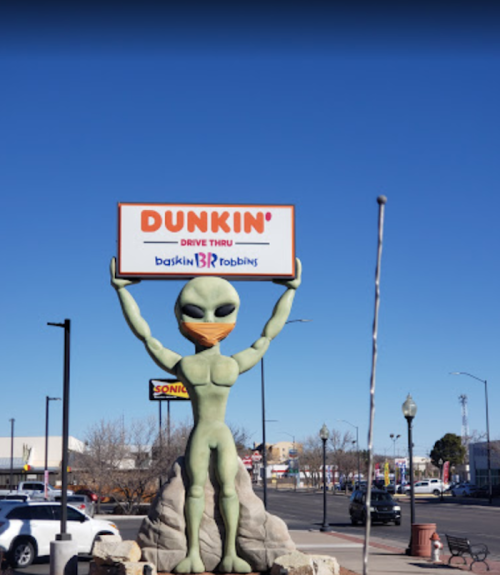 A large green alien statue holds a sign for Dunkin' and Baskin-Robbins in a sunny urban setting.
