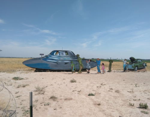 A blue UFO on a dirt road with people and soldiers nearby, alongside an old green car under a clear sky.